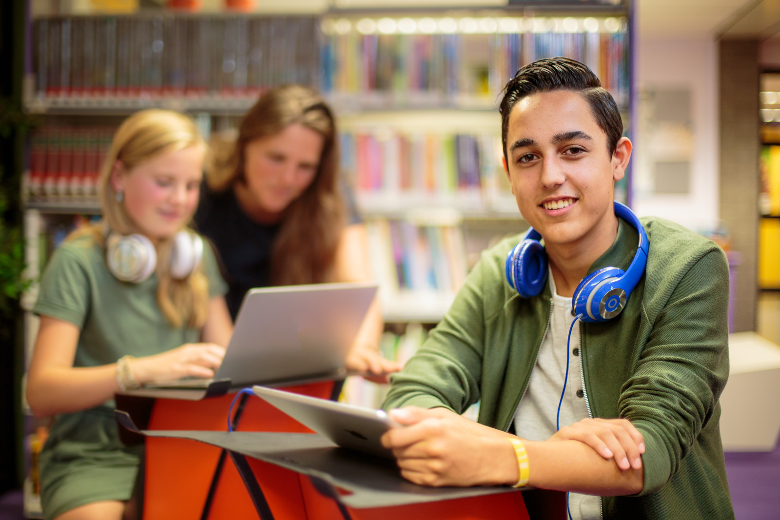 This is a photograph taken in a school library. In the foreground to the right is a teenager sitting at a very small desk with a tablet device in one hand and a set of blue headphones around his neck. He has dark hair and is smiling directly at the camera. Moving backwards, the photograph loses focus but shows a blonde haired girl roughly of ten years old. She has white headphones around her neck and along with a long haired lady sat next to her. Behind    them is an increasingly less focused set of two bookcases. End of description.